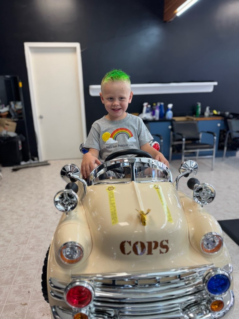 A child with a green mohawk sitting behind the wheel of the kids barber chair